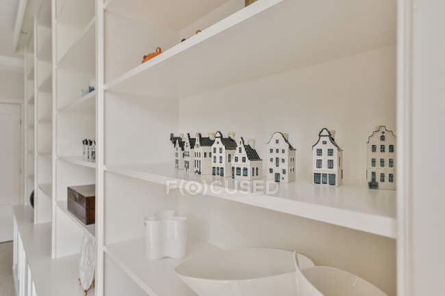 Small ceramic houses arranged in row on shelf of white bookcase in light room — Stock Photo