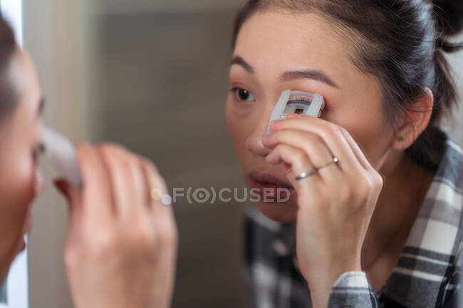 Charming concentrated Asian female using curler on eyelashes while doing makeup in front of mirror at home — Stock Photo