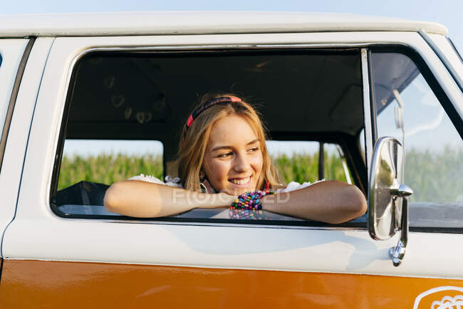 Menina loira bonito sentado dentro de uma van vintage e inclinando-se na janela em um dia ensolarado — Fotografia de Stock