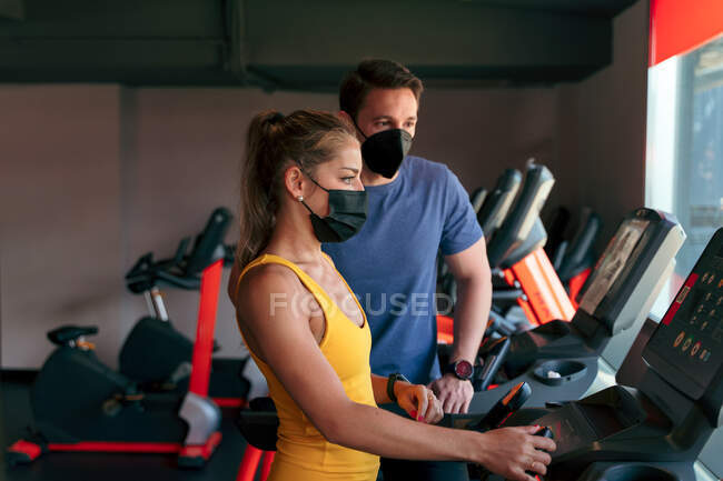Клиентка в защитной маске нажимает кнопки на современной беговой дорожке с цифровым экраном во время тренировки с тренером-мужчиной в спортзале во время вспышки коронавируса — стоковое фото