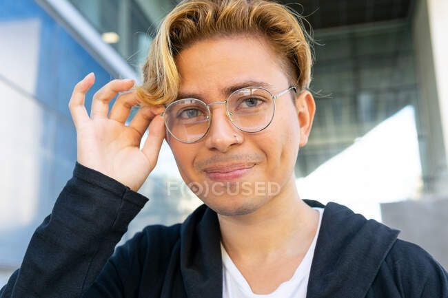 Cheerful young male in casual wear adjusting eyeglasses and looking at camera while sitting on street near modern building in city — Stock Photo