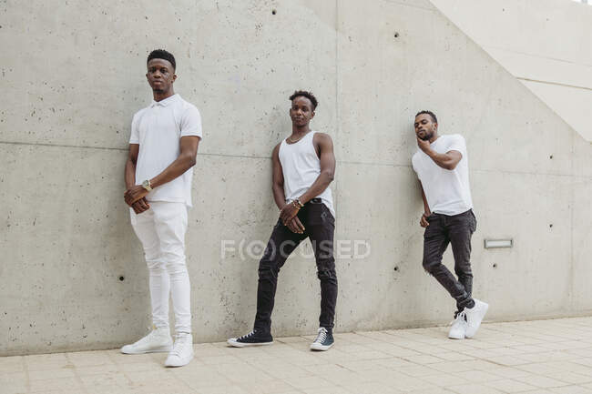 Полное тело крутых афро-американских друзей, одетых в модную одежду с белой футболкой, стоящей рядом со зданием и смотрящих в камеру — стоковое фото