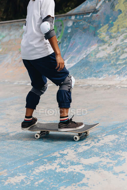 Anonymous young ethnic person in casual outfit wearing protective knee pads riding in skateboard in the skate park — Stock Photo