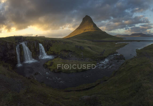 Быстрый каскад, падающий с травянистого холма в реку, текущую рядом с высокой горой против пасмурного неба в природе Исландии — стоковое фото