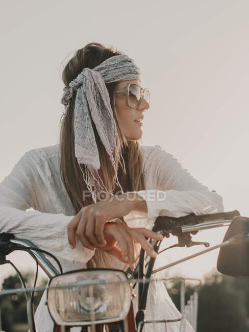 Женщина-хиппи в солнечных очках и платке сидит на мопеде на фоне безоблачного неба на природе во время поездки в солнечное летнее время — стоковое фото