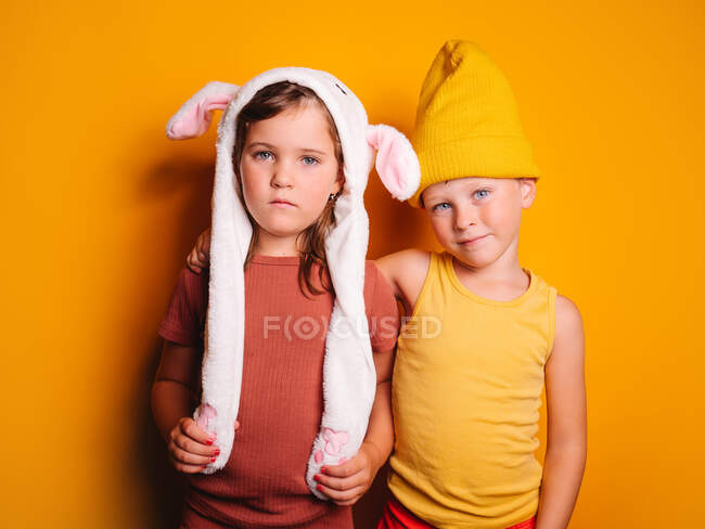 Menino alegre com olhos azuis em camiseta e chapéu de pé e abraçando irmã séria em chapéu branco com orelhas de coelho contra a parede amarela no estúdio — Fotografia de Stock