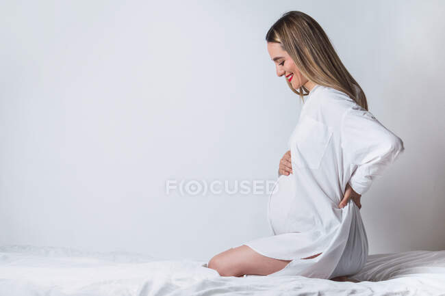 Side view of young gentle female touching tummy while sitting on bed and smiling happily — Stock Photo