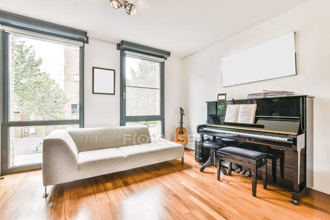 Комфортний диван, розміщений біля чорного піаніно в світлій кімнаті з гітарою і вікнами з видом на вулицю з деревами в стильній квартирі — стокове фото