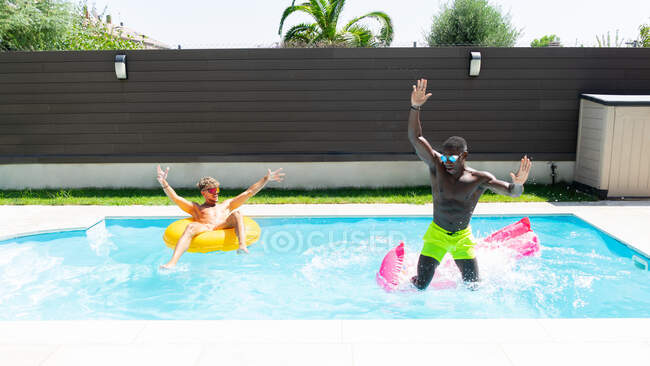 Веселые многорасовые друзья-мужчины веселятся вместе в бассейне, расслабляясь на надувном кольце и матрасе в солнечный летний день — стоковое фото
