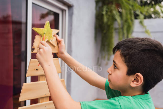 Vista lateral do menino sério com pincel pintura árvore topper de árvore de Natal decorativa com tinta amarela na sala de luz — Fotografia de Stock