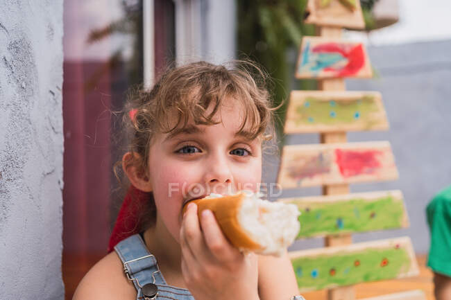 Cute girl looking at camera while eating delicious hot dog in light room with decorative wooden Christmas tree during holiday — Stock Photo