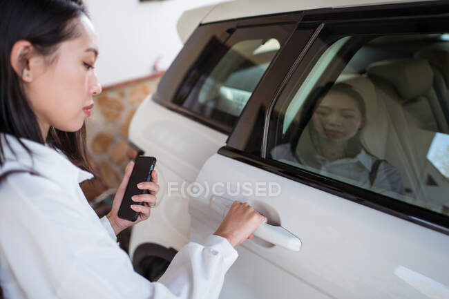 Crop positive ethnic female passenger in formal wear with smartphone opening the back door of the car — Stock Photo