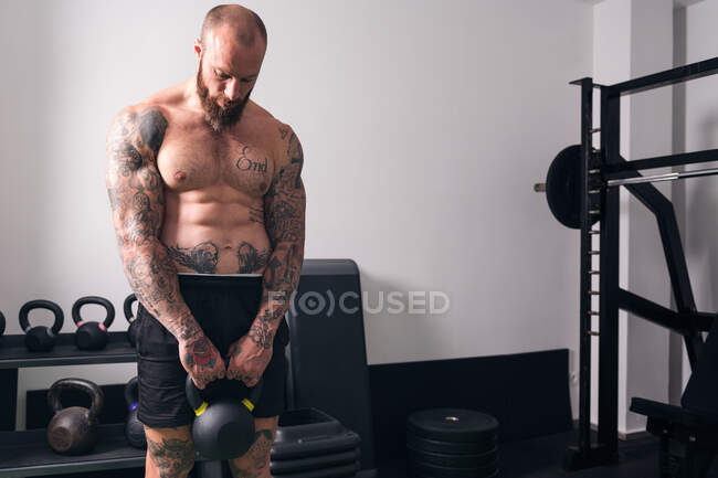 Potente culturista sin camisa con tatuajes haciendo ejercicio con pesadas pesas durante el entrenamiento funcional en el gimnasio - foto de stock