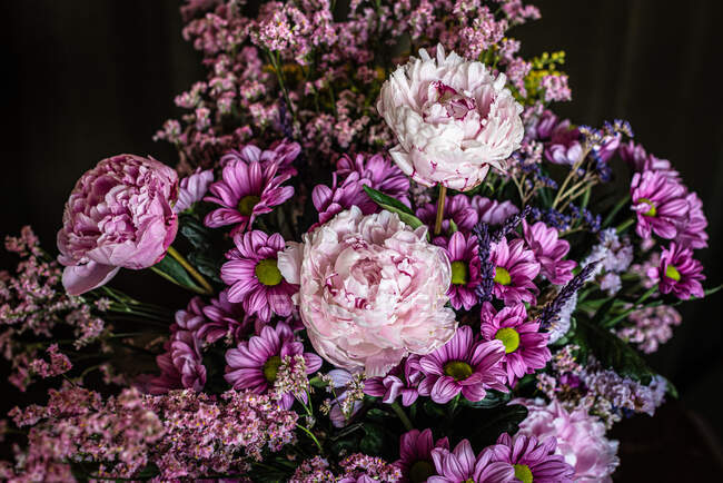 Bouquet de pivoines fraîches colorées et de chrysanthèmes dans un vase en verre posé sur une table en bois dans une pièce sombre — Photo de stock