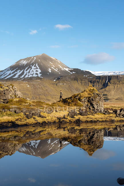 Seitenansicht von jungen männlichen Reisenden in warmer Kleidung, die entlang der steinigen Küste des ruhigen Sees von schneebedeckten Bergen während einer Reise in Island umgeben sind — Stockfoto