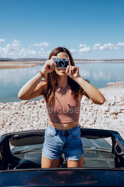 Junge Frau steht auf Auto beim Fotografieren mit altmodischer Fotokamera an der Küste des blauen Teichs — Stockfoto