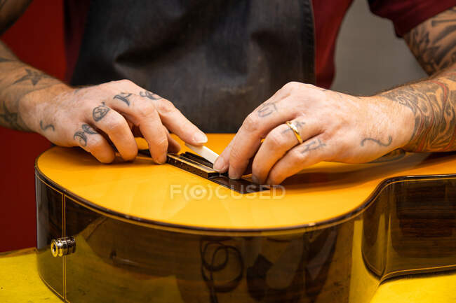 Schnitter anonymer männlicher Geigenbauer mit Tätowierung, weißer Sattel auf Akustikgitarre, während er am Tisch in der leichten professionellen Werkstatt steht — Stockfoto