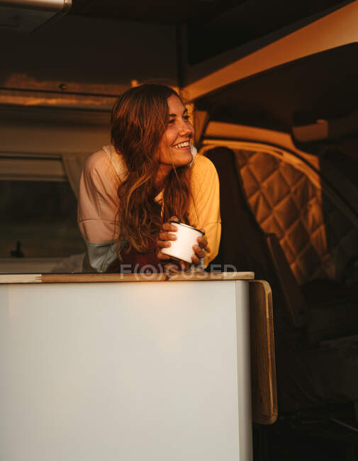 Exploradora femenina encantada con taza de té caliente de pie en campista ambulante al atardecer y disfrutando de la noche en España mientras mira hacia otro lado - foto de stock
