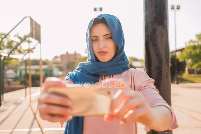 Charming Muslim female in traditional headscarf standing in city street and taking self shot on smartphone on sunny day — Stock Photo