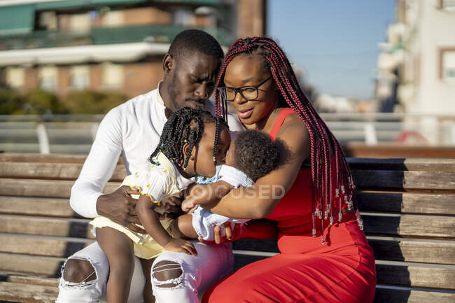Little African American girl sitting on knees of father and kissing baby on hands of mother while sitting together on wooden bench on street in summer day — Stock Photo