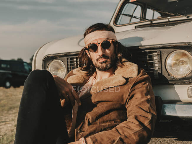 Ernsthafter männlicher Hippie in Oberbekleidung und Stirnband sitzt bei einer Landpartie an sonnigen Tagen in der Nähe eines weißen Oldtimers — Stockfoto