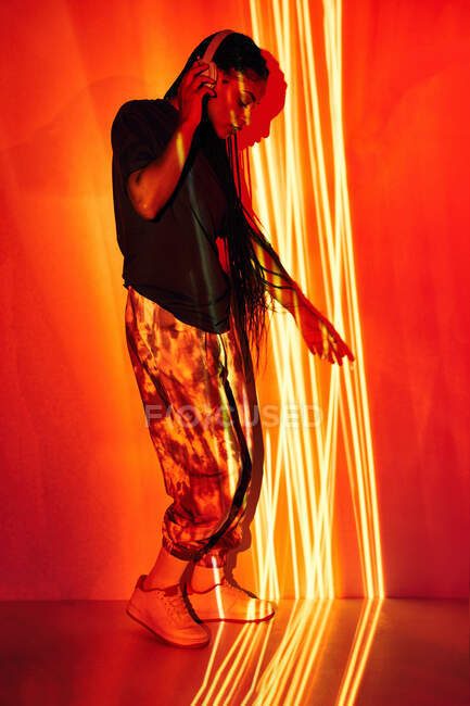 Vue latérale du jeune millénium féminin dominicain à la mode avec de longues tresses afro debout sur le sol et regardant vers le bas tout en écoutant de la musique dans les écouteurs dans la chambre avec un éclairage géométrique coloré — Photo de stock