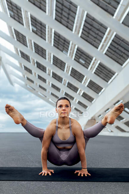 Vue avant du corps complet de la femme forte en vêtements de sport pratiquant la posture Tittibhasana sur le tapis tout en pratiquant le yoga sur la rue avec panneau solaire — Photo de stock