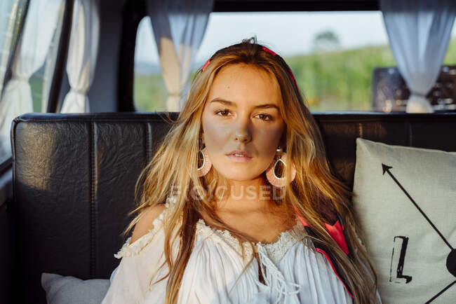 Jolie fille blonde à l'intérieur d'un van vintage et couchée sur le siège par une journée ensoleillée en regardant la caméra — Photo de stock