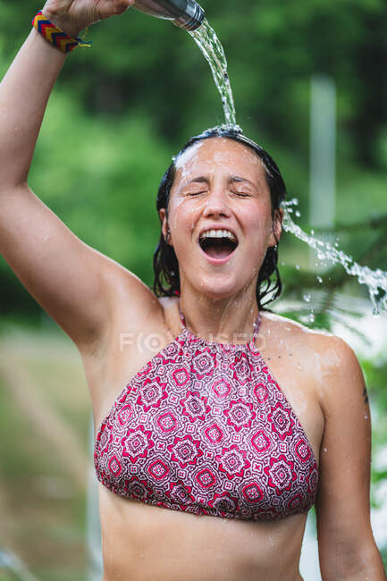 Happy female standing with closed eyes in top and pouring water from bottle on head against green trees in park in summer time — Stock Photo