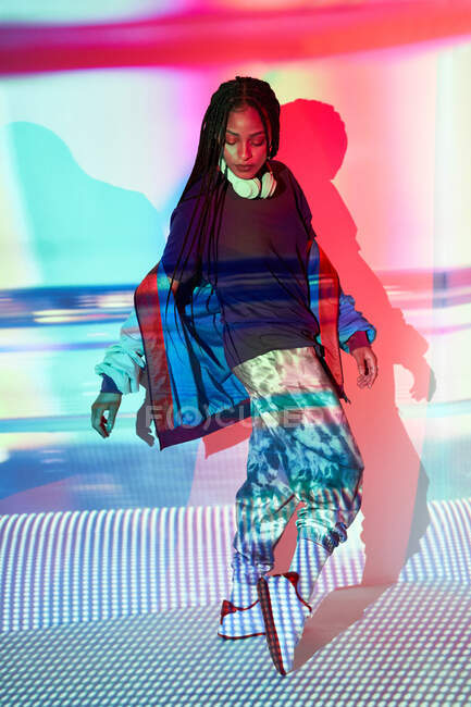 Full body of confident young ethnic female millennial in fashionable outfit and headphones standing on tiptoes near wall with colorful projection and looking down — Stock Photo