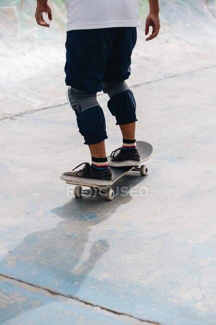 Persona étnica joven anónima en traje casual con rodilleras protectoras montadas en monopatín en el parque de skate - foto de stock