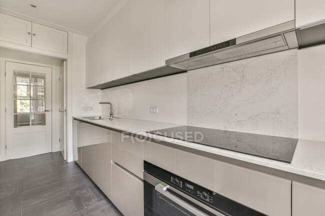 Stretta cucina illuminata dal sole con mobili in stile minimalista bianco e porta balcone in appartamento contemporaneo — Foto stock