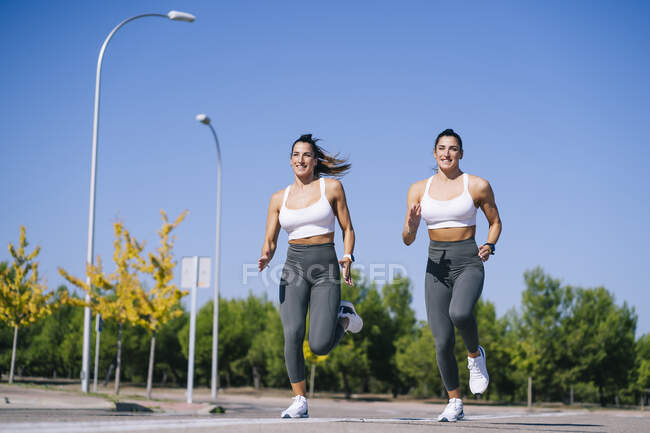 Cuerpo completo de gemelas deportivas sonrientes en ropa deportiva que corren juntas en el camino de asfalto durante el entrenamiento de fitness contra árboles verdes - foto de stock