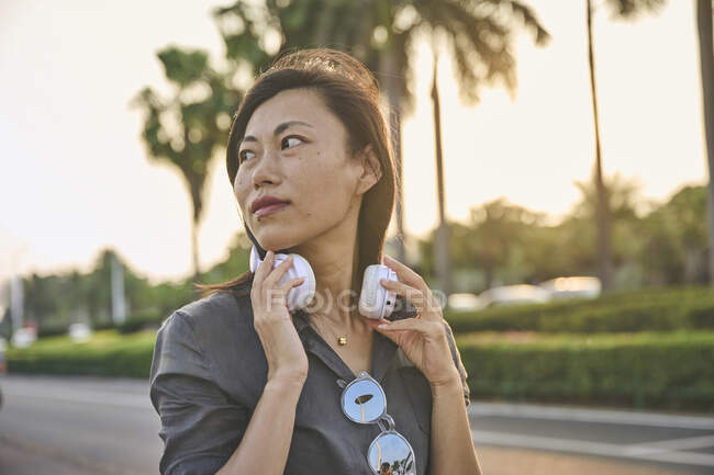 Grave femmina asiatica con moderne cuffie bianche guardando in lontananza mentre in piedi vicino alla strada sulla strada della città con alberi verdi — Foto stock