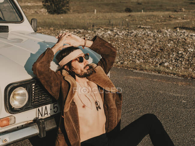 Hippie masculino grave em outerwear e headband sentado perto de automóvel branco velho temporizador durante a viagem no campo no dia ensolarado — Fotografia de Stock
