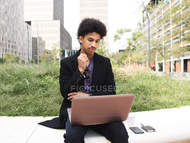 Nachdenklicher junger, selbstständiger Afroamerikaner mit lockigem, dunklem Haar in trendigen Klamotten sitzt im Stadtpark und arbeitet per Laptop an einem Projekt — Stockfoto