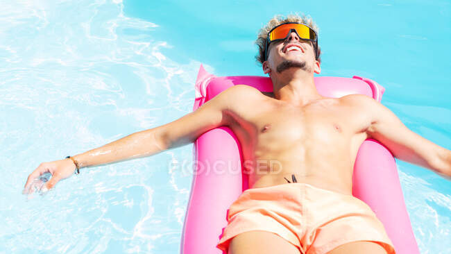 Hombre tranquilo en pantalones cortos de natación y gafas de sol tomando el sol en el colchón inflable rosa en la piscina con agua clara en el día soleado del verano - foto de stock