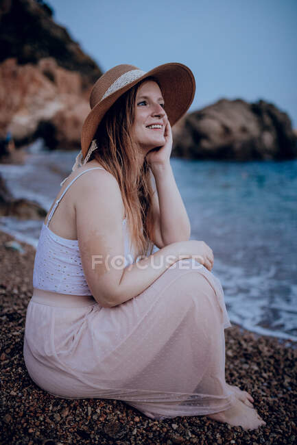 Charming female in hat sitting on beach near sea and touching face while smiling and looking away in summer — Stock Photo