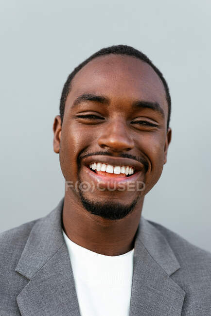 Empreendedor masculino afro-americano sorridente em terno formal sorrindo amplamente enquanto está de pé contra fundo cinza e olhando para a câmera — Fotografia de Stock