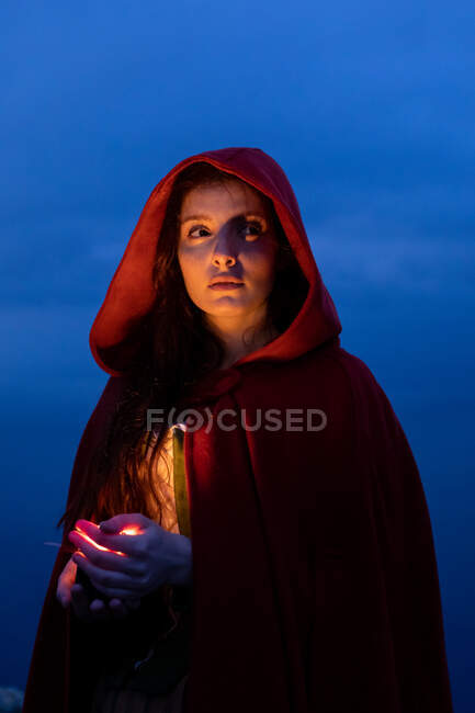 Serious female in Victorian styled outfit with red cape looking away while standing with glowing candle in hands in dark nature — Stock Photo