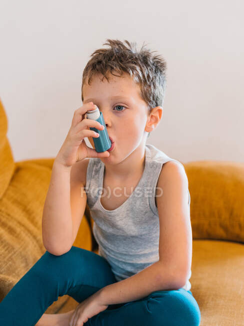 Seitenansicht eines kleinen Jungen mit Asthma beim Inhalieren, während er zu Hause auf dem Sofa sitzt — Stockfoto