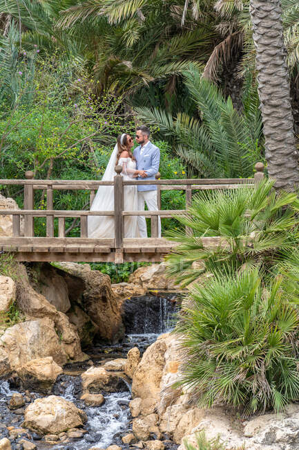 Newlywed couple in wedding outfits standing on wooden footbridge with railing and holding hands while hugging and looking at each other above waterfall with rocks near green trees in park — Stock Photo