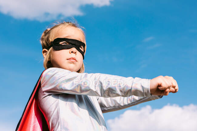 Von unten reckt kleines Mädchen im Superheldenkostüm die ausgestreckten Fäuste, um Macht zu zeigen, während es vor blauem Himmel steht — Stockfoto