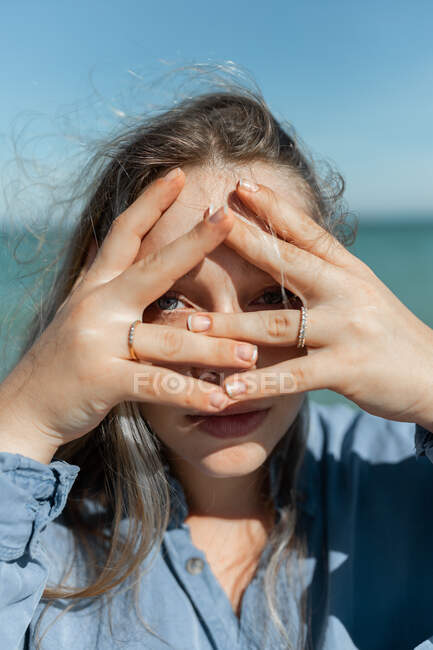 Mujer joven cubriendo la cara con las manos mientras mira la cámara a través de los dedos en la costa en el día soleado de verano - foto de stock