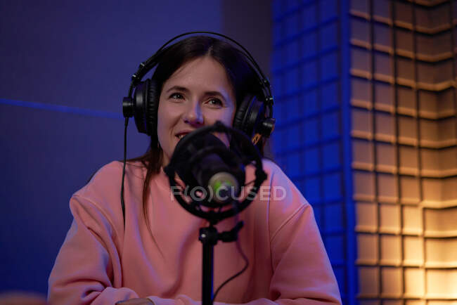 Positive young female radio host in casual clothes and headphones and looking away while recording podcast with male coworker in dark studio — Stock Photo