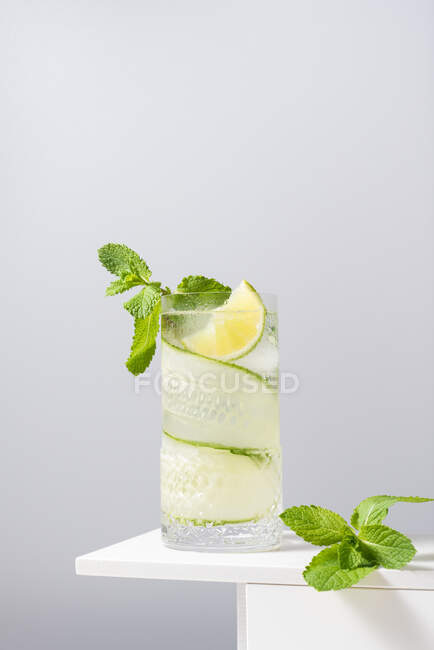 Ein Glas erfrischenden Gin Tonic mit Gurken und Limetten verziert mit Minzblättern auf einem weißen Tisch vor grauem Hintergrund — Stockfoto