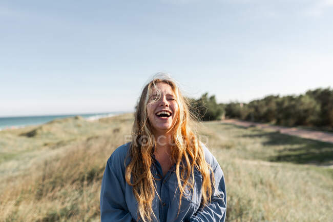 Joven mujer alegre en traje casual de pie en el prado herboso en verano con los ojos cerrados - foto de stock