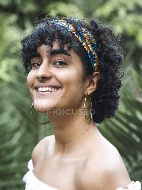 Cheerful charming ethnic female in headband on curly hair looking at camera in green summer garden — Stock Photo