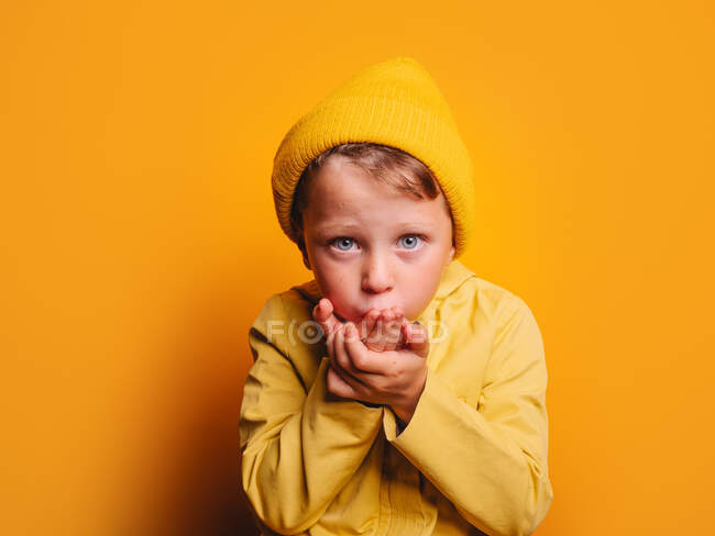 Calm little boy with blue eyes in bright yellow jacket and hat sending air kiss and looking at camera against yellow background in studio — Stock Photo
