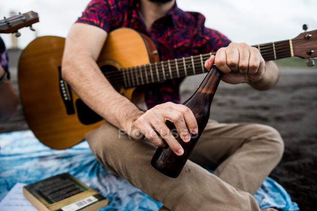 Von unten sieht man einen unkenntlich gemachten männlichen Musiker, der mit Akustikgitarre sitzt und eine Flasche Bier öffnet, während er bei Tageslicht die Zeit am Strand in der Natur verbringt. — Stockfoto